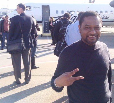 Man smiling and making a peace sign in front of an airplane with passengers in the background.