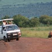 Take trip with us A white van driving on a dirt road with passengers, surrounded by greenery.